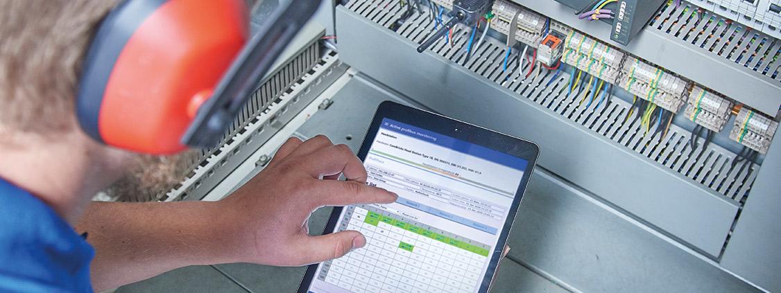 Technician using a tablet to diagnose a PROFIBUS system inside an industrial electrical cabinet.