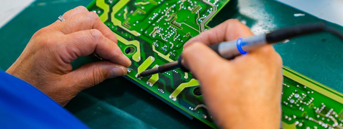 Technician soldering components on a green industrial circuit board during an electronics repair.