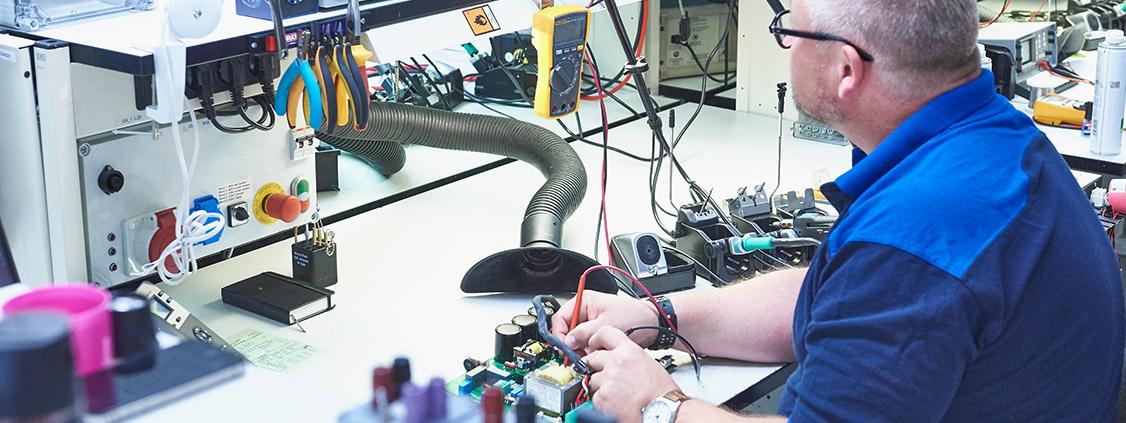 Technician testing an electronic circuit board at a repair workbench using diagnostic equipment.