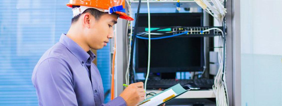 Technician wearing a hard hat inspecting network equipment and recording information on a clipboard.